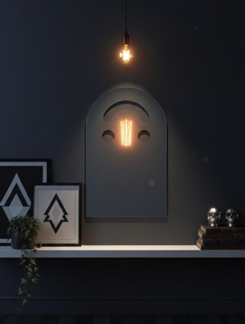 Dark (black) smiley face acrylic mirror, 17.5" tall, hanging on a dark wall above a white floating shelf. A glowing Edison bulb is reflected in the mirror's cut-out. The shelf holds two framed prints with geometric designs, a green plant, and two silver skulls on books. Perfect for gothic bedroom decor.