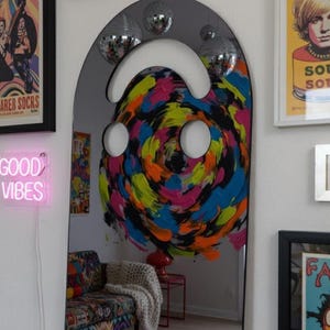 Dark (black) smiley face acrylic mirror, 17.5" tall, hanging on a white gallery wall with other colorful art and disco balls. The mirror reflects a vibrant, swirling abstract painting in pink, blue, yellow, and black, along with a neon "GOOD VIBES" sign. This funky wall art is ideal for eclectic and aesthetic bedroom decor.