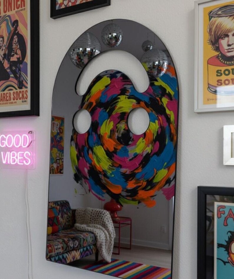 Dark (black) smiley face acrylic mirror, 17.5" tall, hanging on a white gallery wall with other colorful art and disco balls. The mirror reflects a vibrant, swirling abstract painting in pink, blue, yellow, and black, along with a neon "GOOD VIBES" sign. This funky wall art is ideal for eclectic and aesthetic bedroom decor.