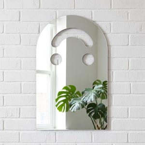 Silver smiley face acrylic mirror, 17.5" tall, hanging on a white brick wall. It reflects a window on the left and a large, dark green monstera plant on the right. The bright silver mirror has silvery undertones, perfect for art pop room decor or alternative style.