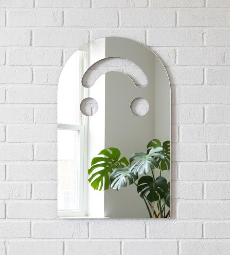 Silver smiley face acrylic mirror, 17.5" tall, hanging on a white brick wall. It reflects a window on the left and a large, dark green monstera plant on the right. The bright silver mirror has silvery undertones, perfect for art pop room decor or alternative style.