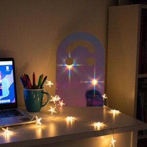 Holo (rainbow) smiley face acrylic mirror, 17.5" tall, standing on a white desk at night. It reflects glowing star fairy lights draped across the desk, casting colorful light flares. Next to it is an open laptop with vibrant wallpaper and a mug of pens. This aesthetic mirror creates a mystical ambiance for a bedroom or dorm.