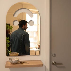 Gold smiley face acrylic mirror, 17.5" tall, hanging by a grey door in a white entryway. It reflects a person in a denim jacket, seen from the back, looking into the mirror. Below is a wooden shelf with keys and two ceramic bowls, perfect as unique aesthetic home decor.