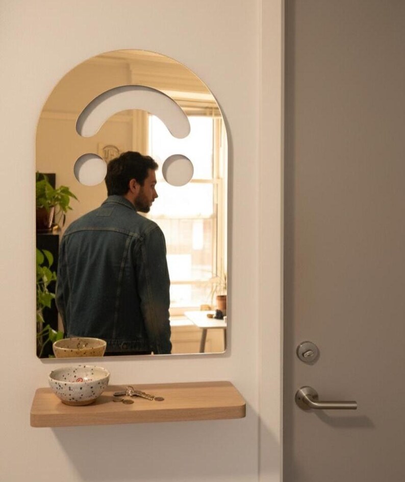 Gold smiley face acrylic mirror, 17.5" tall, hanging by a grey door in a white entryway. It reflects a person in a denim jacket, seen from the back, looking into the mirror. Below is a wooden shelf with keys and two ceramic bowls, perfect as unique aesthetic home decor.