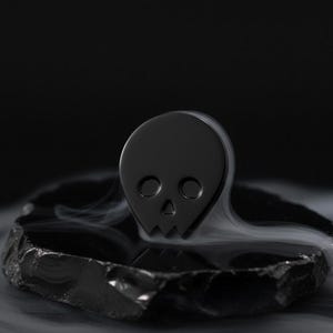 Black minimalist human skull enamel pin (approximately 1 inch tall) sitting on a piece of dark, polished obsidian or slate. Subtle, ethereal wisps of white smoke or mist surround the base of the pin, creating a mystical and contemplative atmosphere. This hero shot emphasizes the "beautiful expanse of the mind" and "goth aesthetic" of the black skull.