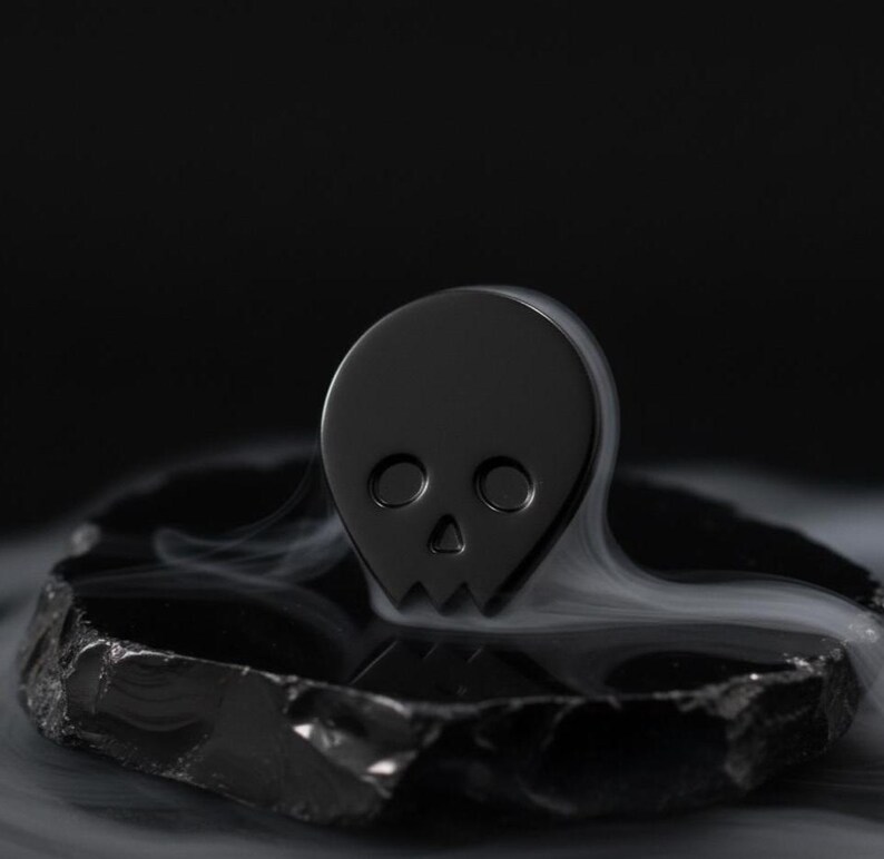 Black minimalist human skull enamel pin (approximately 1 inch tall) sitting on a piece of dark, polished obsidian or slate. Subtle, ethereal wisps of white smoke or mist surround the base of the pin, creating a mystical and contemplative atmosphere. This hero shot emphasizes the "beautiful expanse of the mind" and "goth aesthetic" of the black skull.