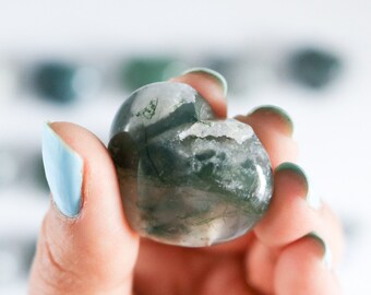 May include: A green and white heart-shaped moss agate crystal. The crystal is being held in a hand with light blue nail polish.