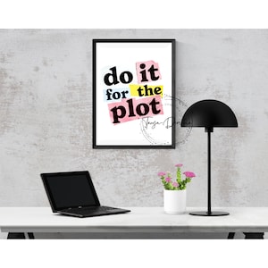 May include: A framed print with the words "do it for the plot" in a colorful, layered design. The print hangs above a white desk with a laptop, a black desk lamp, and a small white pot with pink flowers.