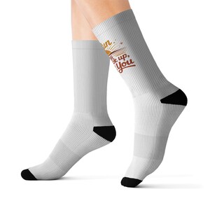 Puede incluir: Un par de calcetines blancos de caña media con talones y punteras negras. Los calcetines presentan un diseño gráfico con un sol estilizado y el texto "Sun is back up, for you" en letras marrones y naranjas en el lateral exterior.