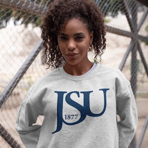 May include: A light gray sweatshirt with the letters "JSU" in navy blue, with "1877" beneath. The sweatshirt is worn by a person with curly dark hair, standing in front of a chain-link fence.