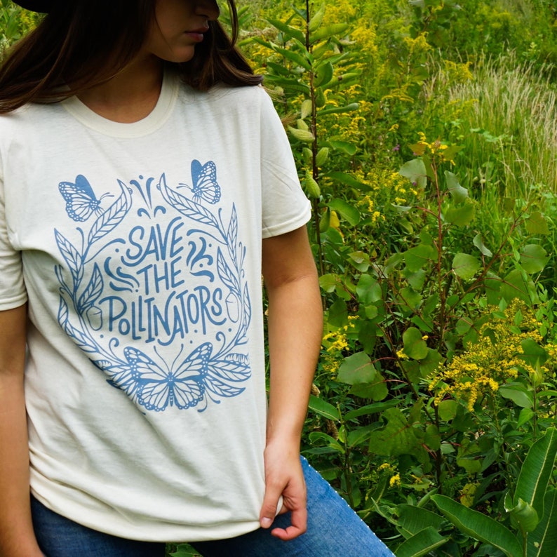 Pu&ograve; includere: Una maglietta bianca con un design grafico blu con farfalle e il testo "Save the Pollinators".