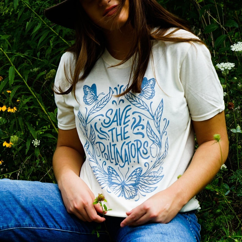 A blue or white shirt with a beautiful custom illustration of the life cycle of the Monarch butterfly. Shows 3 butterflies, milkweed, caterpillars & chrysalis.