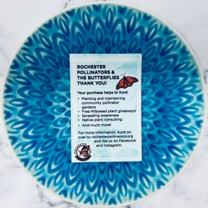 Peut inclure: Une assiette en c&eacute;ramique bleue et blanche avec un motif de papillon. Une carte avec le texte "Rochester Pollinators & The Butterflies Thank You!" est pos&eacute;e sur l'assiette. La carte &eacute;num&egrave;re les moyens de soutenir l'organisation, notamment la plantation de jardins de pollinisateurs, la distribution de plants d'ascl&eacute;piade et la sensibilisation aux plantes indig&egrave;nes.