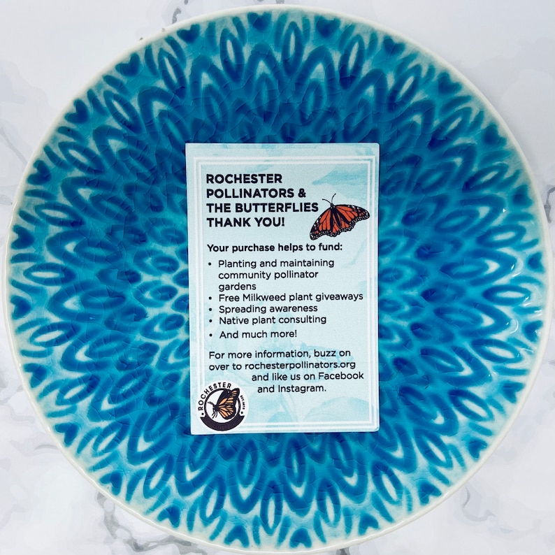 Peut inclure: Une assiette en c&eacute;ramique bleue et blanche avec un motif de papillon. Une carte avec le texte "Rochester Pollinators & The Butterflies Thank You!" est pos&eacute;e sur l'assiette. La carte &eacute;num&egrave;re les moyens de soutenir l'organisation, notamment la plantation de jardins de pollinisateurs, la distribution de plants d'ascl&eacute;piade et la sensibilisation aux plantes indig&egrave;nes.