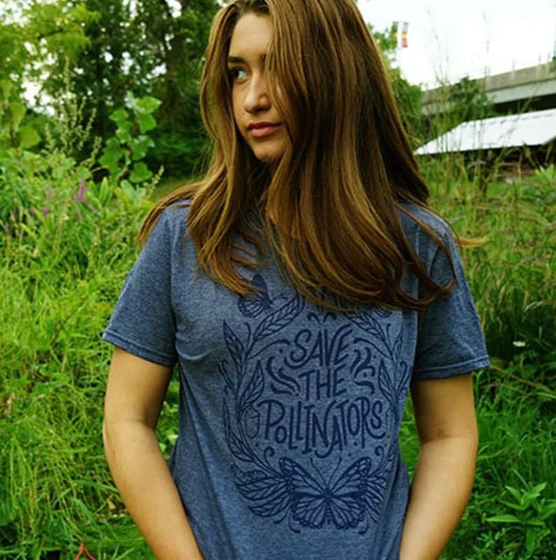 Pu&ograve; includere: Una donna indossa una maglietta blu con un design grafico che dice "Save the Pollinators" con un'illustrazione di farfalla. La maglietta &egrave; realizzata in un materiale morbido e confortevole.
