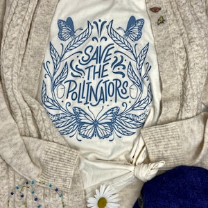 Pu&ograve; includere: Un cardigan beige con una maglietta bianca sotto. La maglietta ha un design grafico blu di farfalle e foglie con il testo "Save the Pollinators".