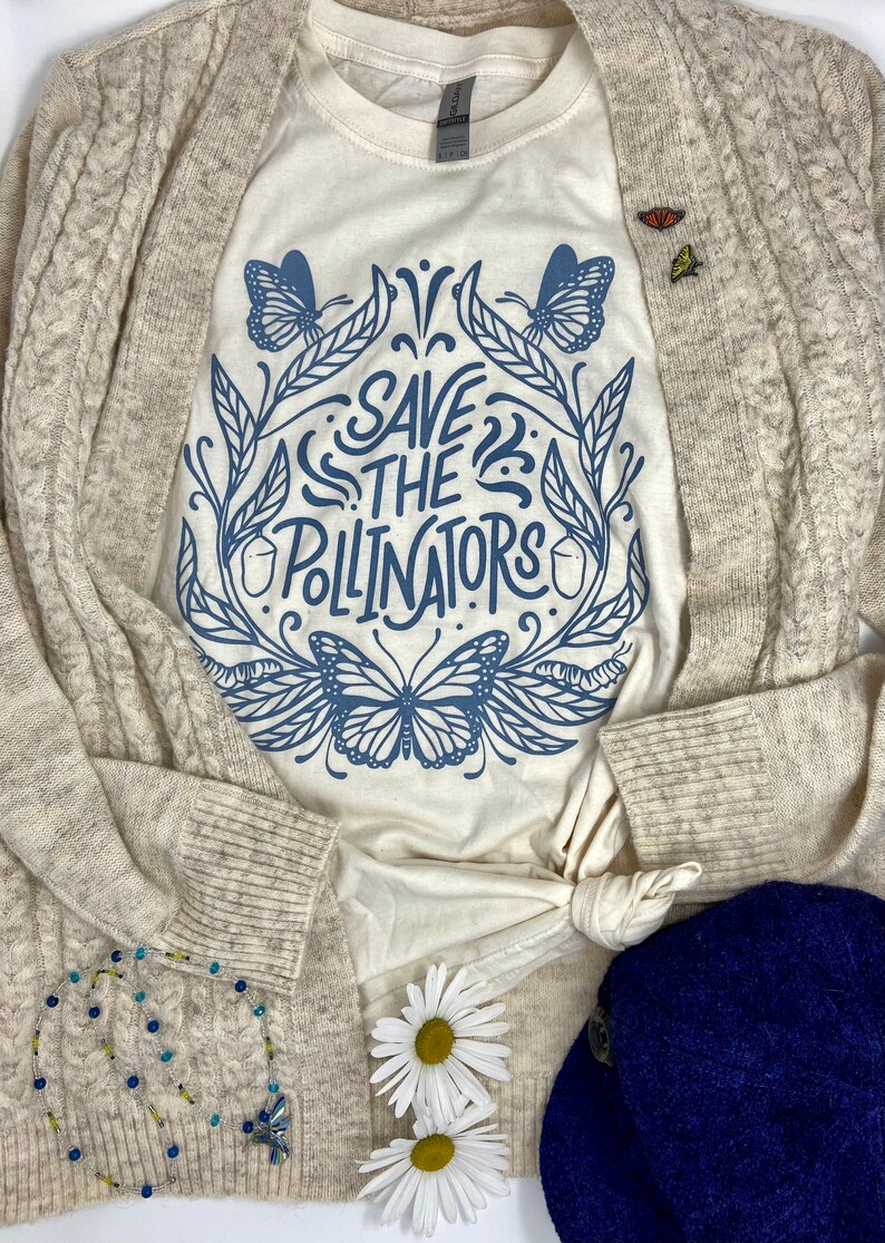 Pu&ograve; includere: Un cardigan beige con una maglietta bianca sotto. La maglietta ha un design grafico blu di farfalle e foglie con il testo "Save the Pollinators".