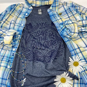 Pu&ograve; includere: Una maglietta grigio scuro con un design grafico blu che dice "Save the Pollinators" con un design di farfalla e fiori. La maglietta &egrave; sovrapposta a una camicia a quadri blu e gialla con un piccolo spillo a farfalla giallo.