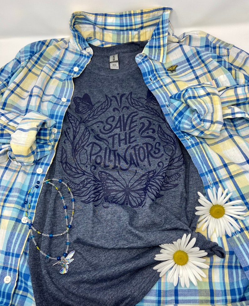 Pu&ograve; includere: Una maglietta grigio scuro con un design grafico blu che dice "Save the Pollinators" con un design di farfalla e fiori. La maglietta &egrave; sovrapposta a una camicia a quadri blu e gialla con un piccolo spillo a farfalla giallo.