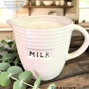 May include: A white ceramic milk pitcher with a ribbed design and a handle. The word "MILK" is printed in black on the front. The pitcher is filled with a white liquid and sits on a wooden surface. The words "Lancaster Mercantile" and "Farmhouse Pitcher" are also visible.