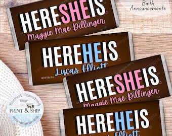 May include: Four chocolate bar birth announcements with the text "HERE SHE IS" and "HERE HE IS" in white, pink, and blue. The names "Maggie Mae Dillinger" and "Lucas Elliott" are printed on the bars. The bars are on a wooden surface.