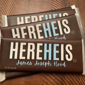 May include: Three chocolate bars with the text "HEREHEIS" in white and light blue. The name "James Joseph Hood" is written below the main text in a cursive font. The bars are wrapped in brown packaging with silver foil ends. Each bar weighs 8 pounds.