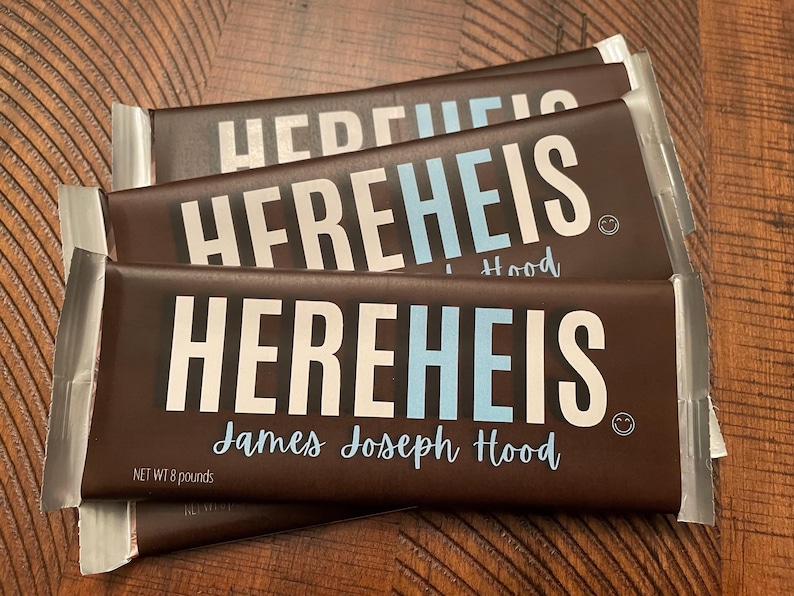 May include: Three chocolate bars with the text "HEREHEIS" in white and light blue. The name "James Joseph Hood" is written below the main text in a cursive font. The bars are wrapped in brown packaging with silver foil ends. Each bar weighs 8 pounds.