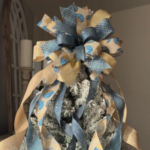 May include: A Christmas tree topper featuring a large bow made of blue, gold, and tan ribbons. The bow has a plaid pattern, glitter accents, and a pattern of blue circles. Long ribbons cascade down the tree, creating a festive holiday decoration.