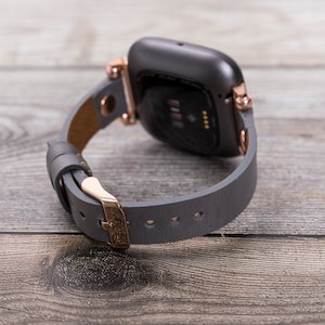May include: A gray leather watch band with rose gold hardware. The band is attached to a black smartwatch with a rose gold bezel.