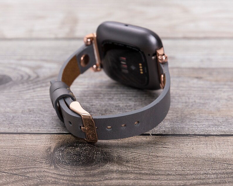 May include: A gray leather watch band with rose gold hardware. The band is attached to a black smartwatch with a rose gold bezel.