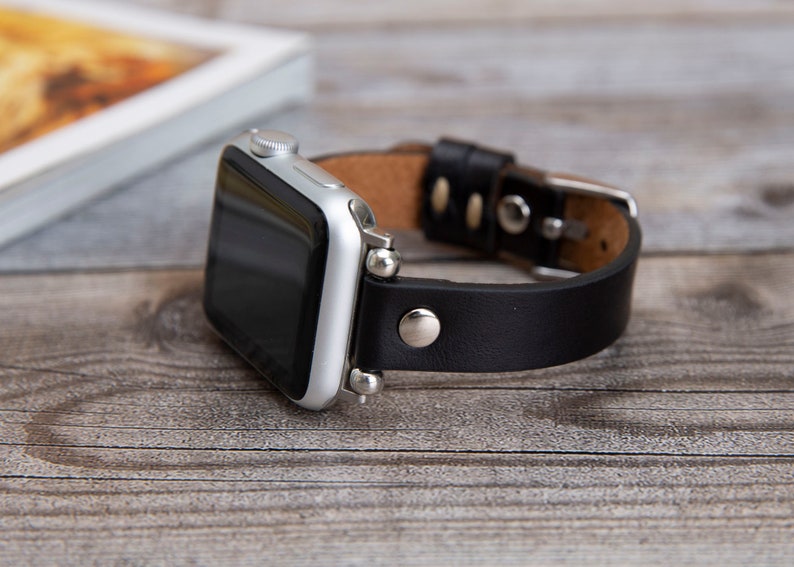 May include: A silver Apple Watch with a black leather band. The watch face is black and the band has a buckle closure.