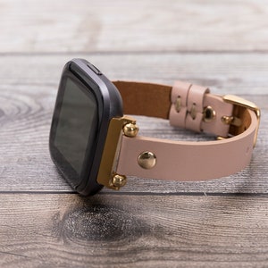 Puede incluir: Un reloj inteligente negro y dorado con una correa de cuero rosa. El reloj tiene una esfera cuadrada y una pantalla digital.