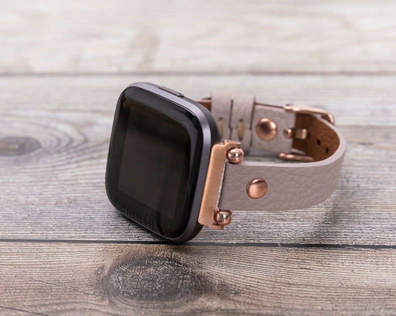Puede incluir: Una correa de reloj de cuero gris con detalles en oro rosa y una esfera de reloj inteligente negra.