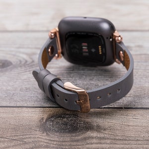 May include: A gray leather watch band with rose gold hardware and a gray smartwatch. The band has a buckle closure.