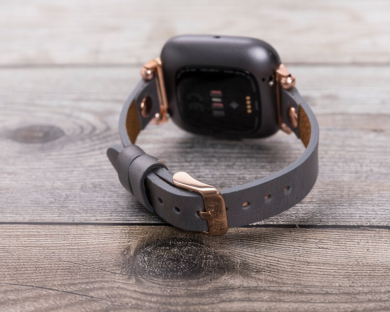May include: A gray leather watch band with rose gold hardware and a gray smartwatch. The band has a buckle closure.