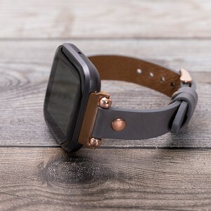 May include: A black smartwatch with a gray leather band and rose gold accents. The band has a buckle closure.