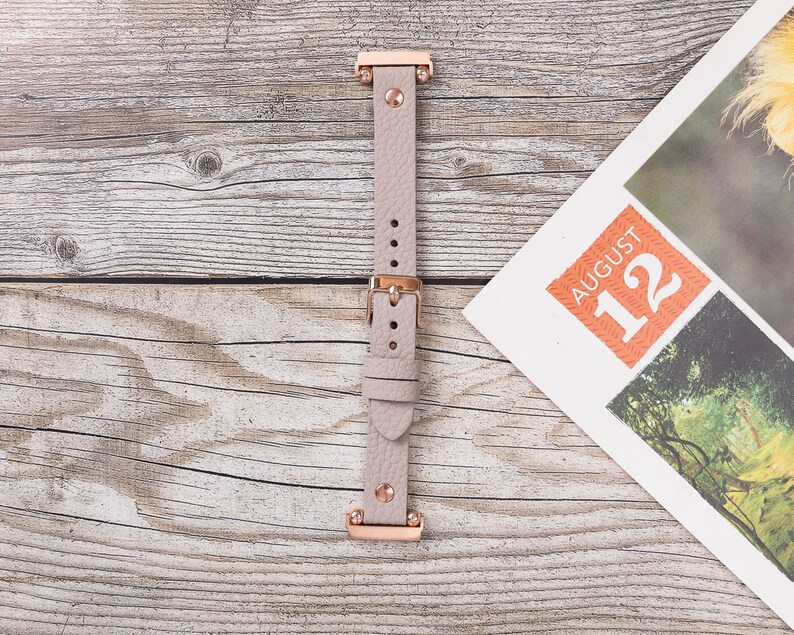 May include: A light pink leather watch band with rose gold hardware. The band has a buckle closure and is shown on a wooden surface.