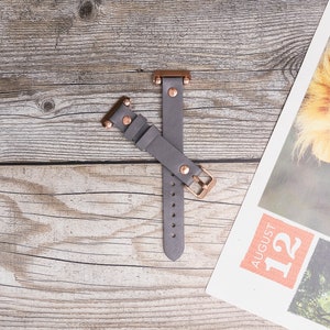 May include: A gray leather watch band with rose gold hardware. The band has a buckle closure and is shown on a wooden surface.