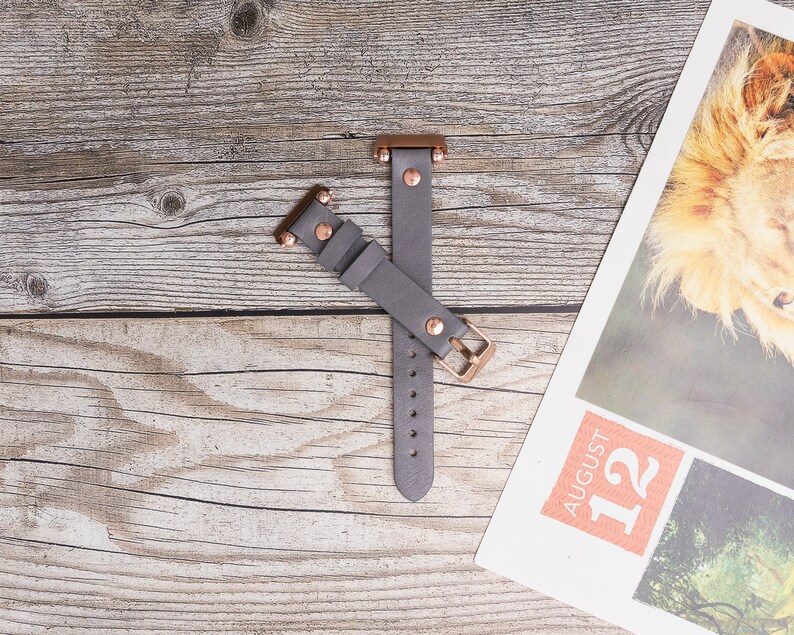 May include: A gray leather watch band with rose gold hardware. The band has a buckle closure and is shown on a wooden surface.