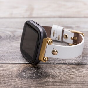 May include: A black smartwatch with a white leather band and gold accents. The band has gold studs and a gold buckle.