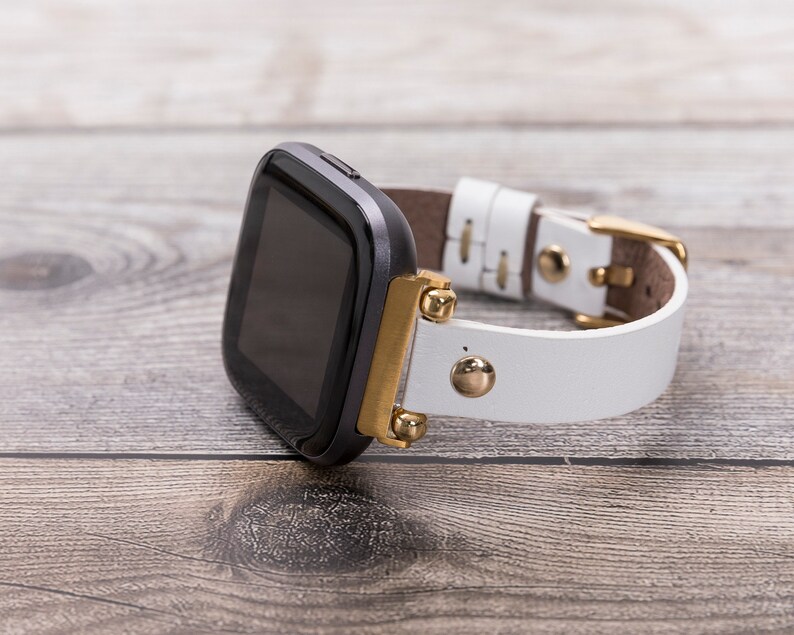 May include: A black smartwatch with a white leather band and gold accents. The band has gold studs and a gold buckle.