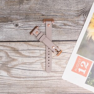 May include: A light gray leather watch band with rose gold accents and a buckle closure.