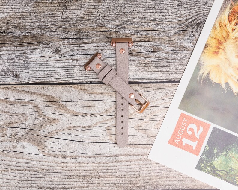 May include: A light gray leather watch band with rose gold accents and a buckle closure.
