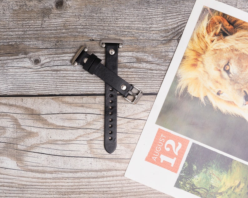 May include: Black leather watch band with silver hardware. The band is on a wooden surface.