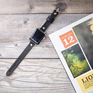 May include: A black leather watch band with silver accents and studs. The watch face is black and the band is on a wooden surface. The background includes a magazine with the text "August 12" and "Lion King".