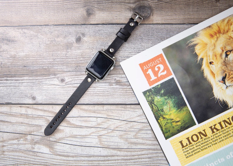 May include: A black leather watch band with silver accents and studs. The watch face is black and the band is on a wooden surface. The background includes a magazine with the text "August 12" and "Lion King".