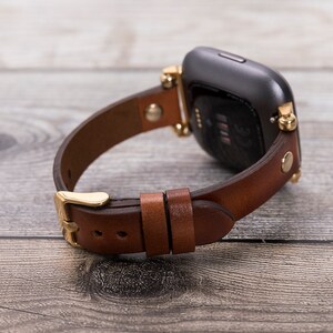 以下が含まれることがあります： 正方形のスマートウォッチ用の、金色の金具とバックルが付いた茶色の革製の腕時計バンドです。