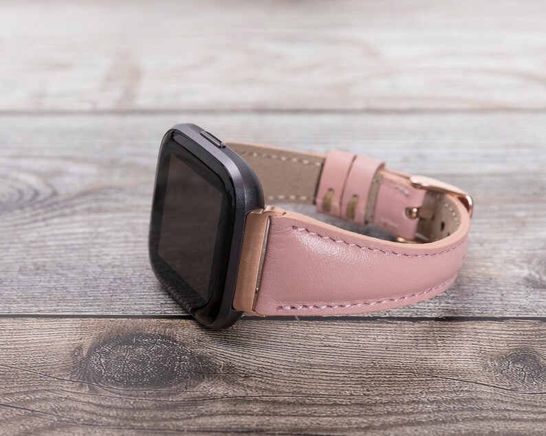 Puede incluir: Una correa de reloj de cuero rosa con hebilla de oro rosa y una esfera de reloj inteligente negra.
