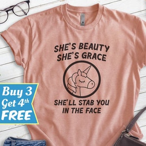 Puede incluir: Camiseta rosa polvoriento con el texto "SHE'S BEAUTY SHE'S GRACE SHE'LL STAB YOU IN THE FACE" y un gr&aacute;fico de unicornio negro. La camiseta se muestra con vaqueros azules y un bolso marr&oacute;n oscuro.