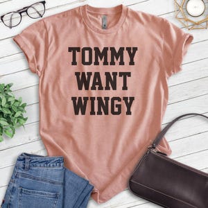 May include: A dusty rose-colored t-shirt with the text "TOMMY WANT WINGY" in bold, dark brown letters. The shirt is folded, with a pair of blue jeans and a black purse in the frame. A pair of glasses and a small plant are also visible.